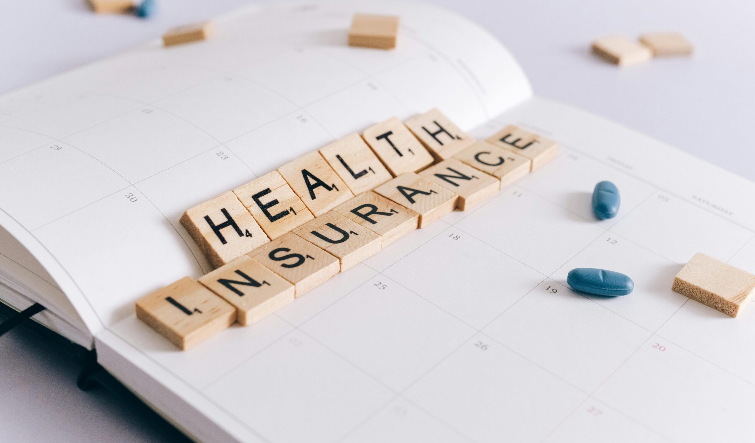 Scrabble tiles spelling 'Health Insurance' on a calendar with pills.